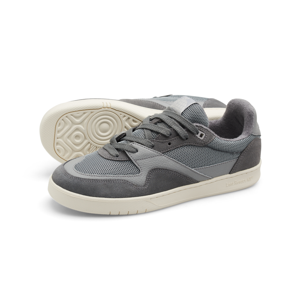 Introducing the LAST RESORT AB CM002 ULTIMATE GREY sneakers by Last Resort AB. These gray shoes feature tactile green mesh panels and white soles, enhanced with suede accents and leather trim. One shoe is seen with its laces tied, while the other leans slightly to the side, capturing a dynamic look.