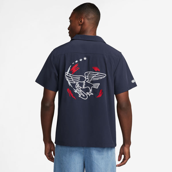 A person wearing the NIKE SB 2024 Federation Kit Button-Up Skate Bowler in USA Obsidian with a white and red winged shoe graphic design on the back, featuring Nike SB's Dri-FIT technology, is facing away from the camera.