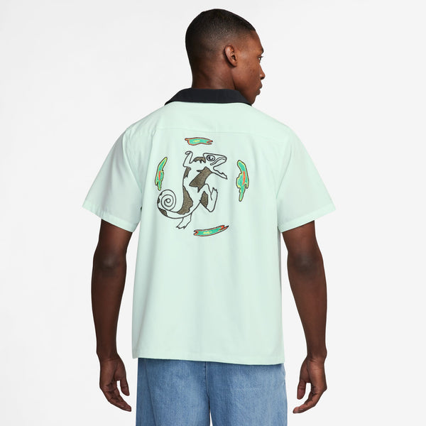 A person is wearing a Nike SB 2024 Federation Kit Button-Up Skate Bowler in Barely Green and Black. This Team USA jersey features colorful feather-like designs surrounding a chameleon graphic on the back and includes a customizable jock tag. The person is facing away.