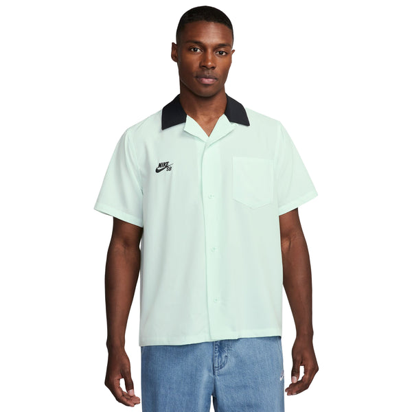 Person wearing a NIKE SB 2024 FEDERATION KIT BUTTON-UP SKATE BOWLER in Barely Green/Black, styled like a short-sleeve button-up shirt with a black collar and a Nike logo, paired with light blue jeans, standing against a white background.