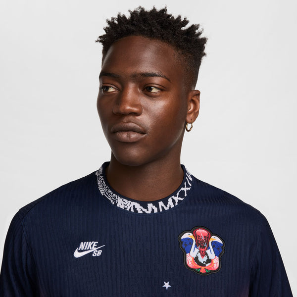 A person wearing a NIKE SB 2024 Federation Kit Dri-FIT ADV short-sleeve skate jersey in USA Obsidian/Bright Crimson/White, featuring a colorful embroidered patch on the chest and a patterned neckline, looking to the side against a plain background.