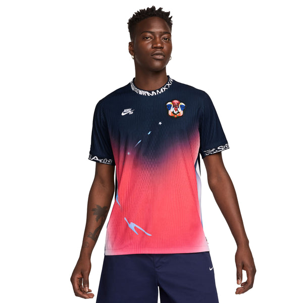 A person wearing a multicolored gradient NIKE SB 2024 Federation Kit Dri-FIT ADV short-sleeve skate jersey in USA Obsidian/Bright Crimson/White with a patch on the chest stands against a white background.