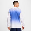 A man stands with his back to the camera, wearing a NIKE SB 2024 Federation Kit long sleeve skate tee in white, showcasing a gradient blue and white design with an American flag on the back, symbolizing global inspiration.