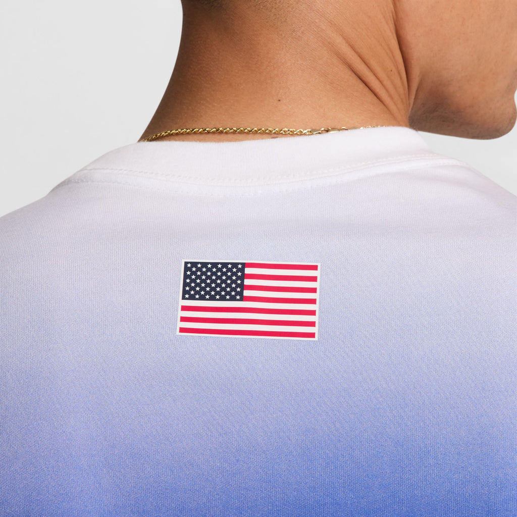 Close-up of the back of a person's neck wearing a Nike SB 2024 Federation Kit long sleeve skate tee, USA white, in midweight cotton. The tee features a white to blue gradient design and a small American flag on the back, complemented by a gold chain around the neck.