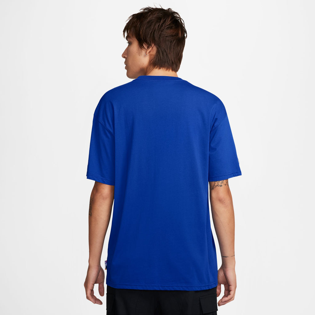 Seen from the back, a person with short hair is wearing a plain blue t-shirt from the NIKE SB 2024 Federation Kit Skate Tee USA Old Royal and black pants against a white background, sporting a NIKE cap.