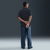 A person stands facing away with hands clasped behind their back, wearing a black and white striped shirt and Nike SB Loose Double-Knee Denim Skate Pants, made from durable denim, against a plain dark background.