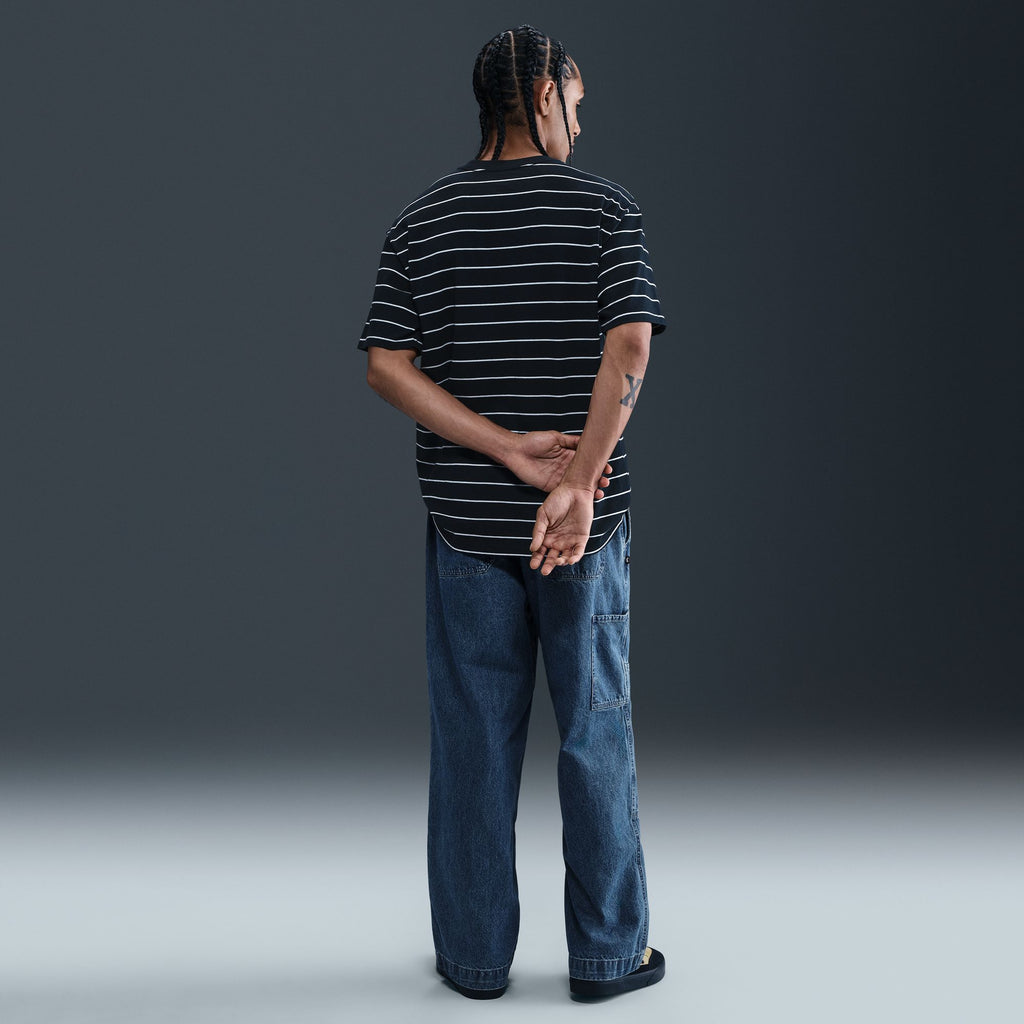 A person stands facing away with hands clasped behind their back, wearing a black and white striped shirt and Nike SB Loose Double-Knee Denim Skate Pants, made from durable denim, against a plain dark background.