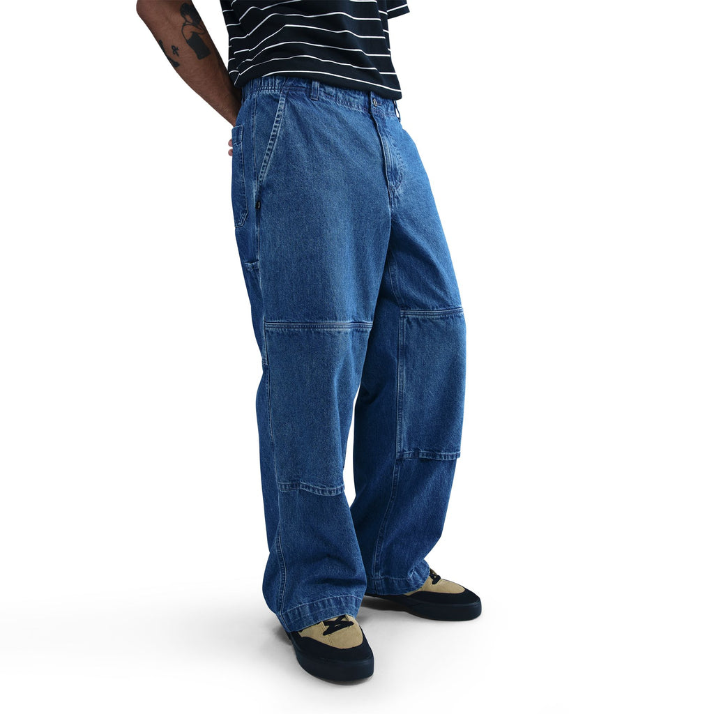 Model wears NIKE SB LOOSE DOUBLE-KNEE DENIM SKATE PANTS by nike—loose blue denim—paired with a black and white striped shirt and black-and-beige shoes, standing against a plain white background.
