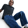 A person with braided hair wears a black and white striped t-shirt and nike SB Loose Double-Knee Denim Skate Pants, sitting on the floor against a plain white background.