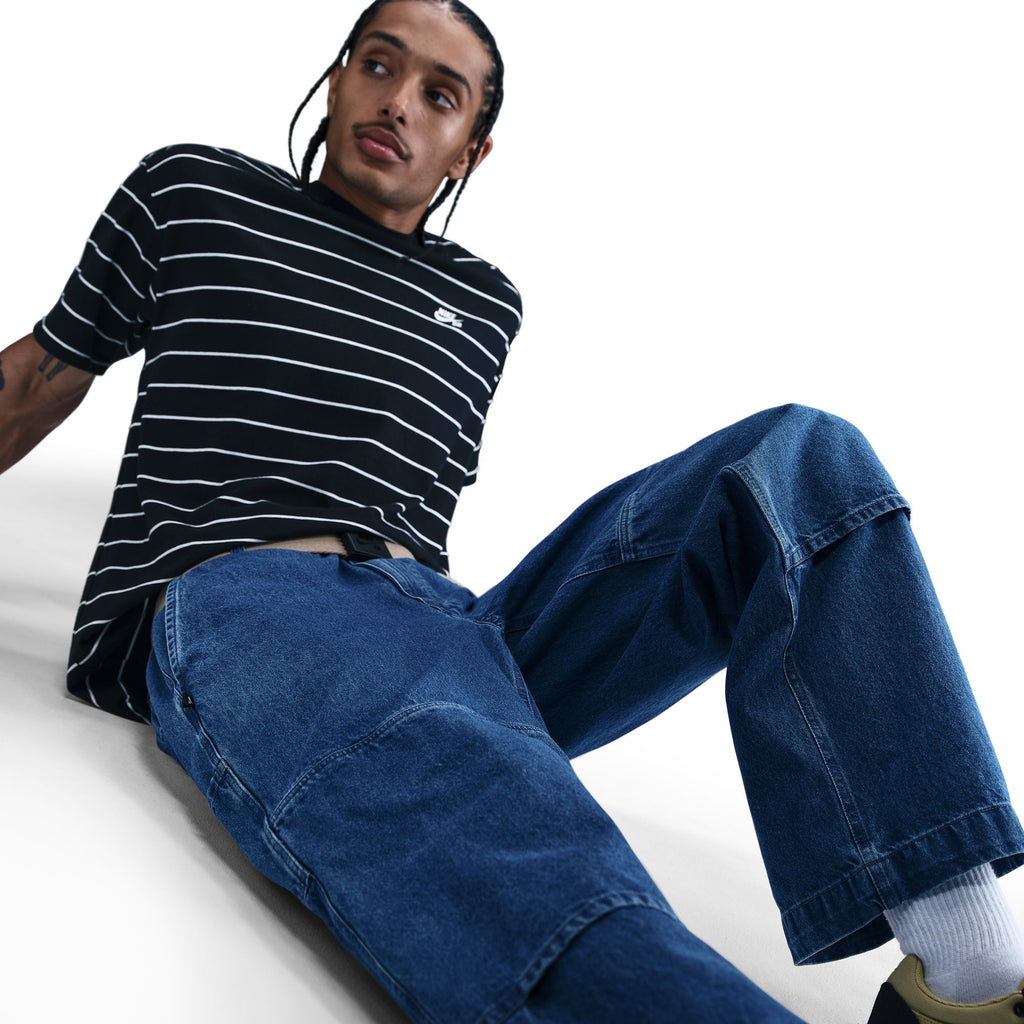 A person with braided hair wears a black and white striped t-shirt and nike SB Loose Double-Knee Denim Skate Pants, sitting on the floor against a plain white background.