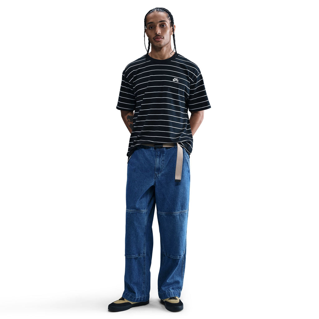 A person stands against a white background wearing a black and white striped t-shirt, nike NIKE SB LOOSE DOUBLE-KNEE DENIM SKATE PANTS, a tan belt, and black shoes.