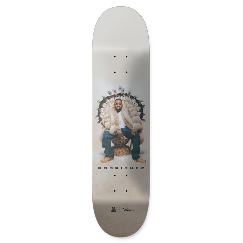 The PRIMITIVE X OUTKAST Rodriguez Regal skateboard deck features a person seated on an ornate chair wearing a fur coat and jeans, with "Rodriguez" elegantly printed below. It evokes the style and culture of an Outkast album cover.