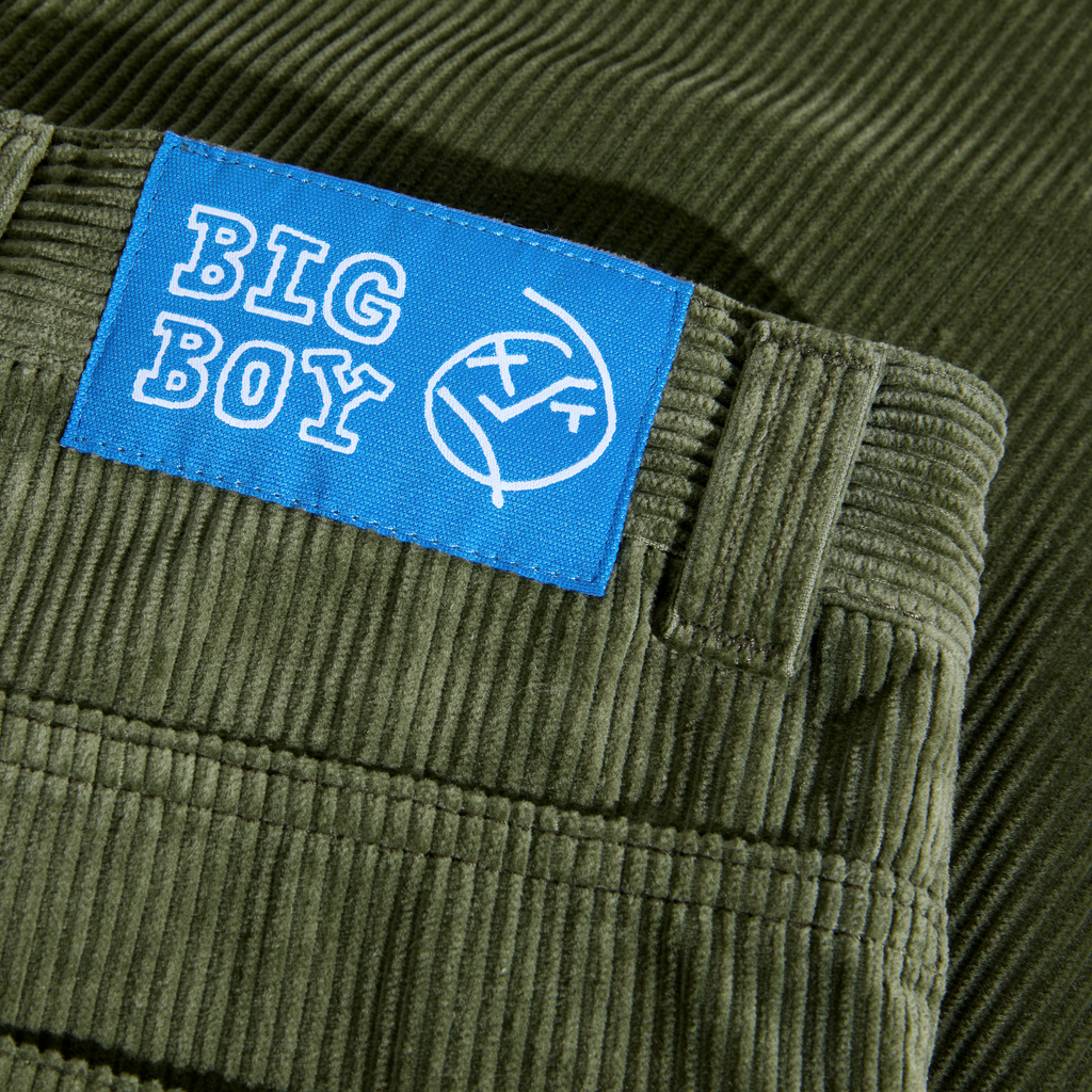 Close-up of POLAR BIG BOY CORD UNIFORM GREEN pants, crafted from green corduroy fabric, with a blue tag displaying the "BIG BOY" label alongside a face graphic, and designed with a baggy fit for enhanced comfort.