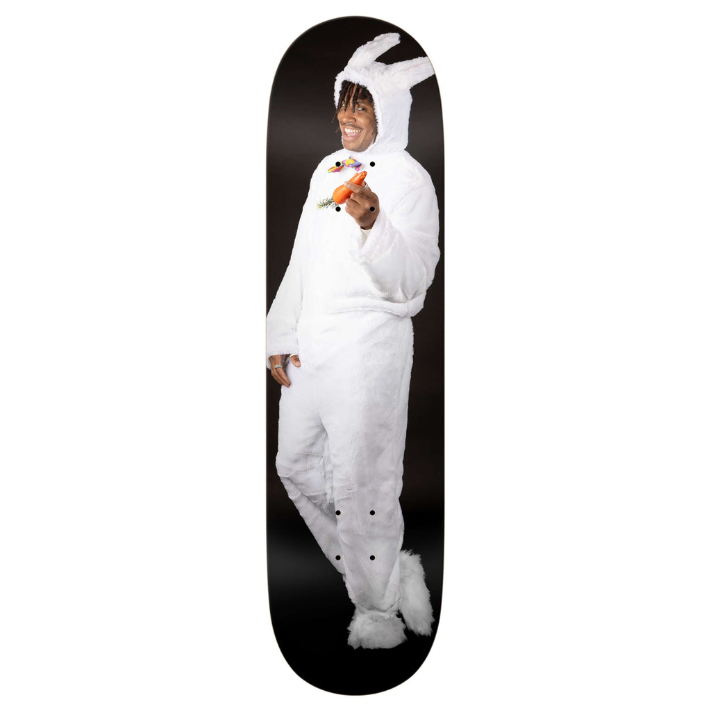 A person in a white rabbit costume holds a carrot and smiles, standing against a black background on the REAL ISHOD BUNNY TWIN TAIL skateboard by REAL.