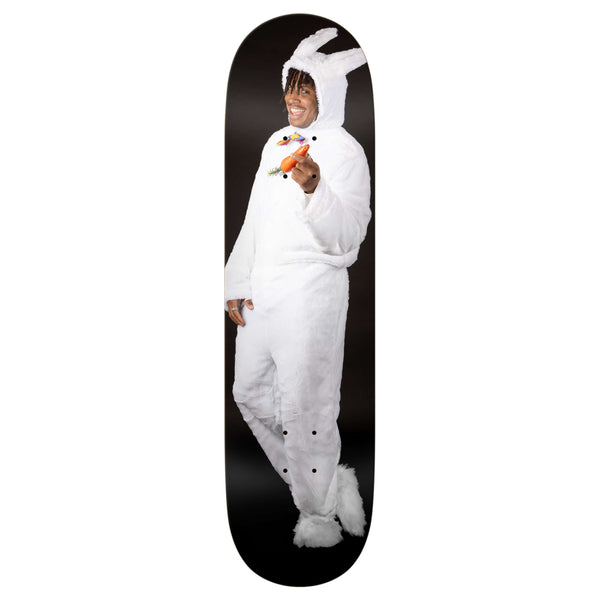 A person in a white rabbit costume holds a carrot and smiles, standing against a black background on the REAL ISHOD BUNNY TWIN TAIL skateboard by REAL.
