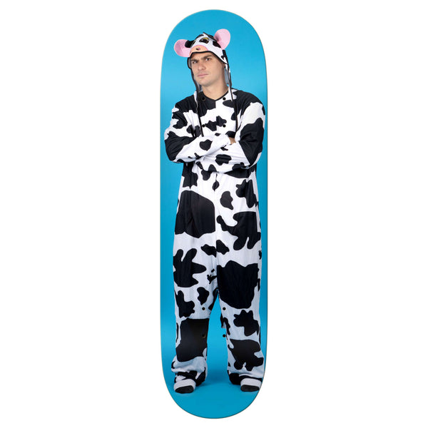 A person stands with arms crossed, wearing a black and white cow costume with a hood featuring ears and pink horns, holding the REAL MASON COW skateboard by REAL against a blue background.