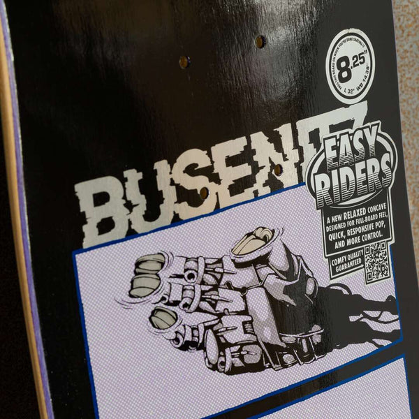 Close-up of a skateboard deck featuring an illustration of a gloved fist and part of the word "BUSENITZ." A sticker for "EASY RIDERS" showcases product specifications, such as its relaxed concave design for better board control and responsive pop. This deck is from the REAL BUSENITZ 4TH WALL EASY RIDERS collection by REAL.