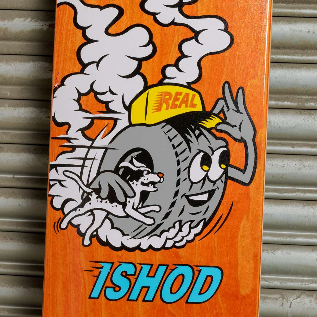 The REAL ISHOD MASCOT skateboard deck features a playful cartoon design of a tire with eyes and arms, sporting a yellow cap, in energetic chase of a spotted dog. Boasting an original shape and an 8.06" width, the word "ISHOD" adorns the bottom, combining style with a sense of fun.