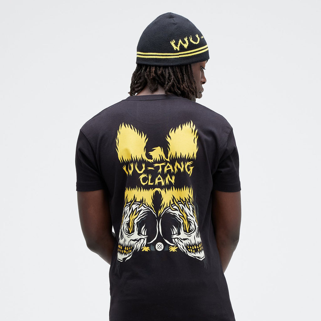 A person wearing the black STANCE X Wu-Tang Skull Clan Tee, crafted from a soft cotton blend, pairs it with a beanie featuring yellow designs and stands facing away.