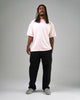 A man with long dreadlocks stands facing forward against a plain gray background, wearing a light pink t-shirt, black pants, and WARSAW LTD's CHAPTER | STONE FG NUBUCK minimalistic footwear.