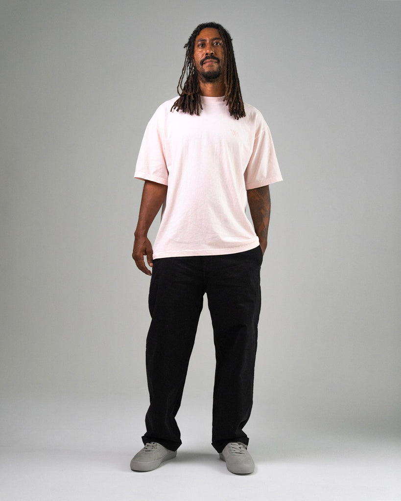 A man with long dreadlocks stands facing forward against a plain gray background, wearing a light pink t-shirt, black pants, and WARSAW LTD's CHAPTER | STONE FG NUBUCK minimalistic footwear.