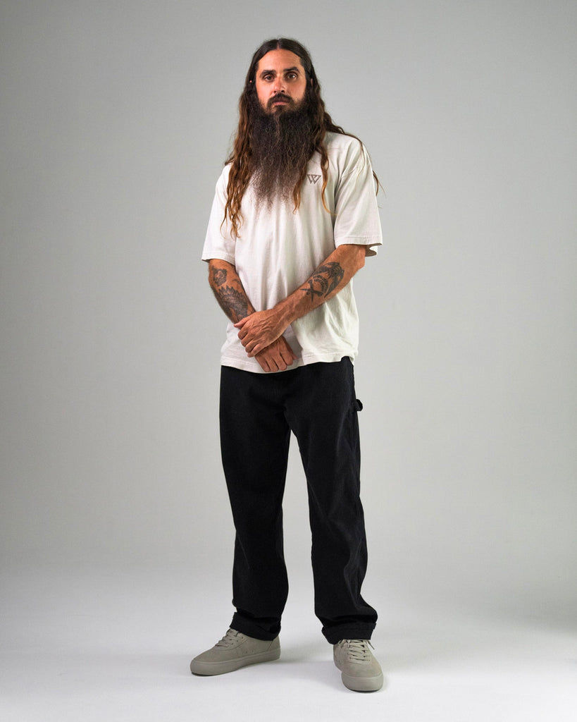 A man with long hair and a beard stands facing forward, wearing a white t-shirt, black pants, and WARSAW LTD CHAPTER MID | STONE FG NUBUCK skate-inspired footwear, with his hands clasped in front.