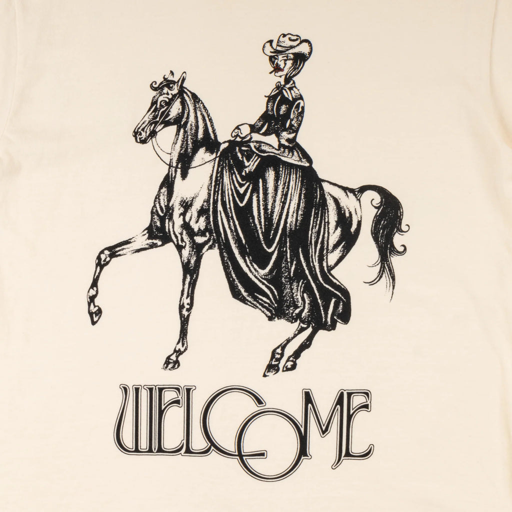 A WELCOME COWGIRL GARMENT-DYED TEE BONE with an image of a woman riding a horse.