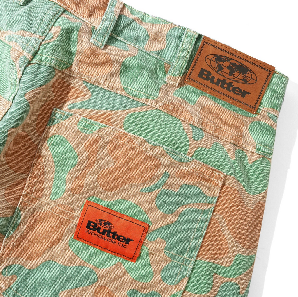 A pair of BUTTER GOODS SANTOSUOSSO CAMO PANTS with an orange logo on the pocket.