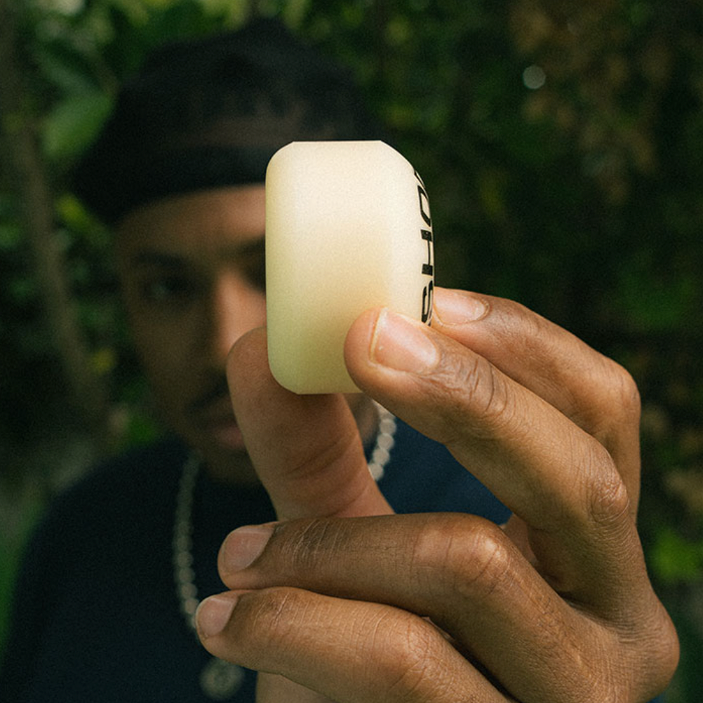 Someone holds a white SPITFIRE F4 ISHOD LOCK-IN CLASSICS 99D 52MM skateboard wheel close to the camera, with blurred greenery in the background.