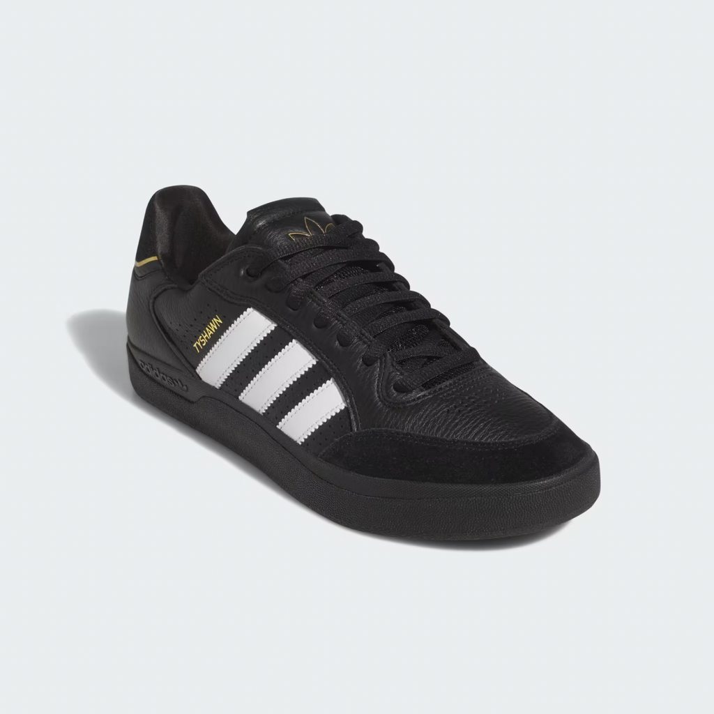 The ADIDAS TYSHAWN LOW in Core Black, Cloud White, and Gold Metallic showcases its design with iconic white stripes and gold text. Enhanced with Adiprene cushioning for skateboarding comfort, it stands out against a light gray background.