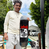 A man in light clothing stands outdoors with a black and white dog, holding the REAL ISHOD VICIOUS TWIN TAIL 8.5" skateboard deck by REAL. Trees, a pool, and cars are visible in the background.