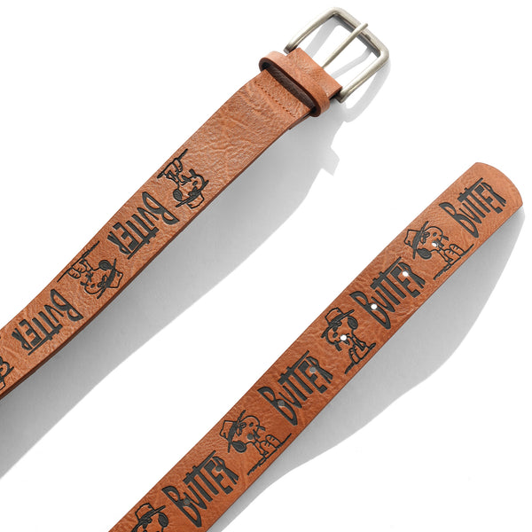 The BUTTER X PEANUTS SPIKE BELT BROWN by Butter Goods is a brown PU leather belt with metal buckle, "BUTTER" and cartoon dog graphics in black, plus an embossed pattern for extra style.