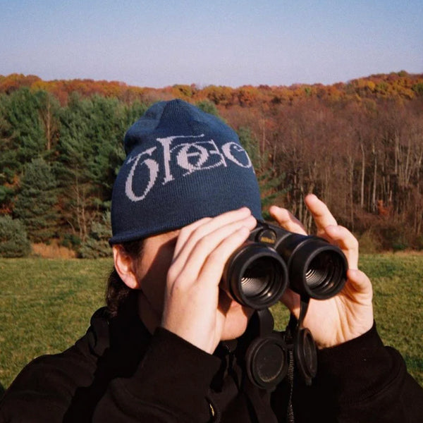 A person wearing a THEORIES SECRETUM JAQUARD KNIT BEANIE MARINE with a logo looks through binoculars in a wooded area.