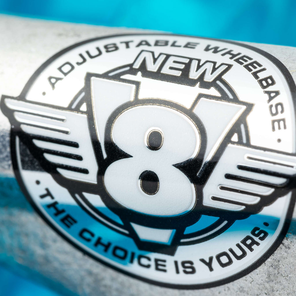 Close-up of a metallic logo reading "NEW V8 ADJUSTABLE WHEELBASE THE CHOICE IS YOURS," spotlighting VENTURE V8 TEAM EDITIONS 5.2 HI POLISHED skateboard trucks, with a blue blurred background emphasizing the innovative VENTURE design.