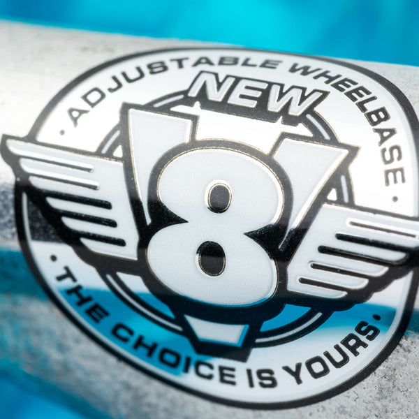 Close-up of a metallic logo reading "NEW V8 ADJUSTABLE WHEELBASE THE CHOICE IS YOURS," spotlighting VENTURE V8 TEAM EDITIONS 5.2 HI POLISHED skateboard trucks, with a blue blurred background emphasizing the innovative VENTURE design.
