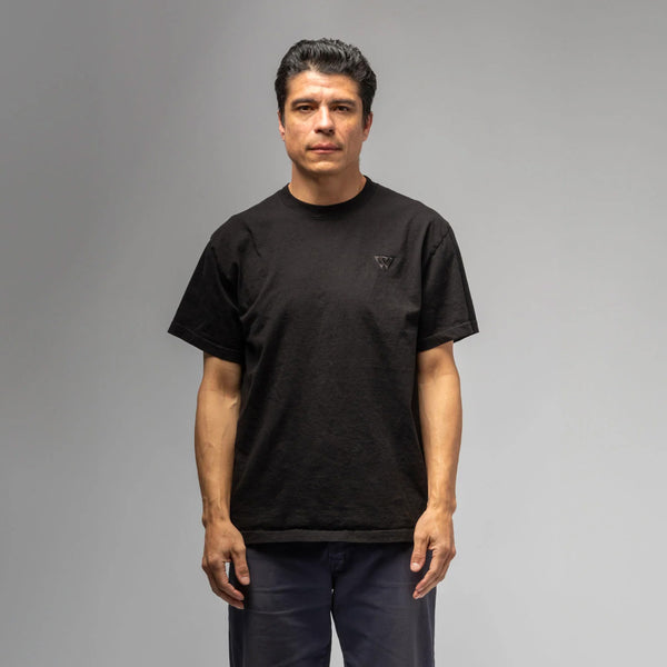 A man with short dark hair is against a gray backdrop, wearing a black WAR SAW UNITY TEE made from 100% cotton and dark pants, looking directly at the camera.