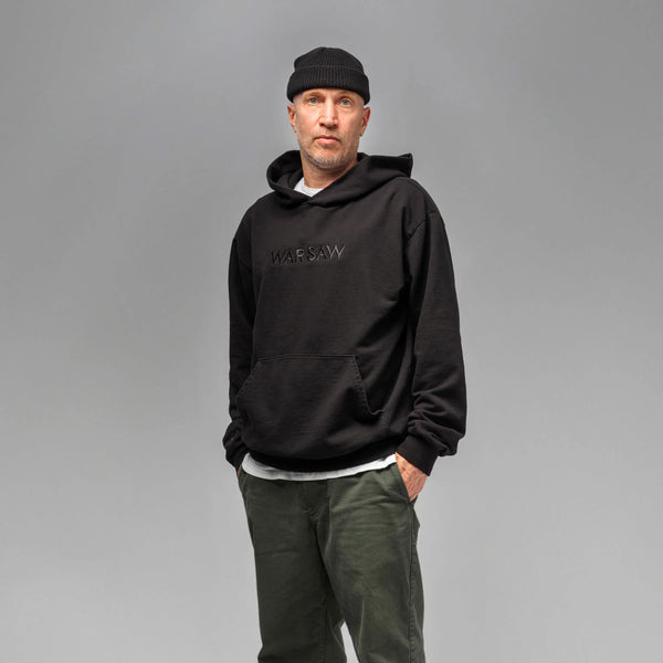 A man in a WAR SAW UNKNOWN HOODIE BLACK with an oversized unisex fit and beanie stands against a gray background, embodying casual style.