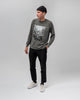 A man wearing a black beanie, grey graphic long-sleeve shirt, black pants, and WARSAW LTD CHAPTER | WHITE / WHITE sneakers stands against a plain light grey background.