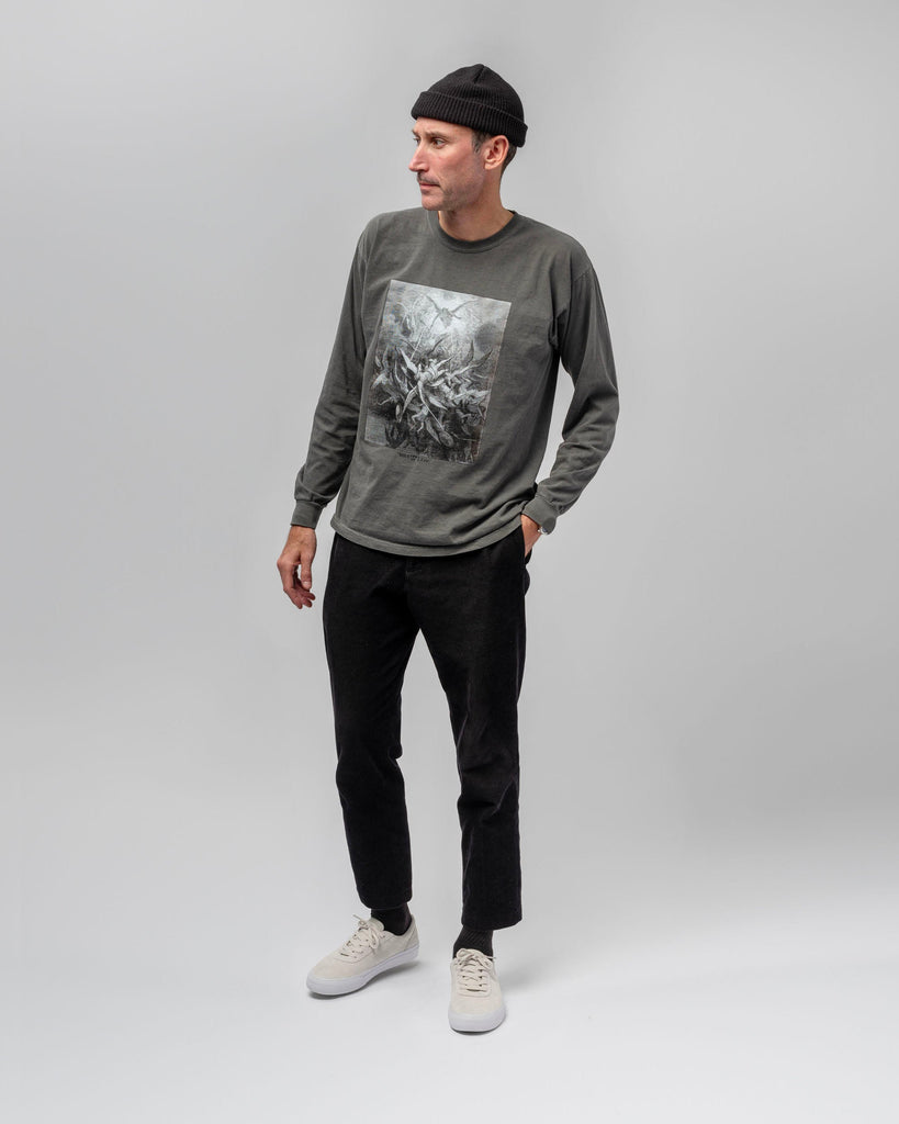 A man wearing a black beanie, grey graphic long-sleeve shirt, black pants, and WARSAW LTD CHAPTER | WHITE / WHITE sneakers stands against a plain light grey background.