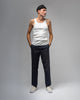 A man in a black beanie, white tank top, dark pants, and WARSAW LTD's STRANGER SLIP-ON | WHITE / WHITE stands with hands in pockets against a plain gray background, showcasing comfort and durability in his understated style.