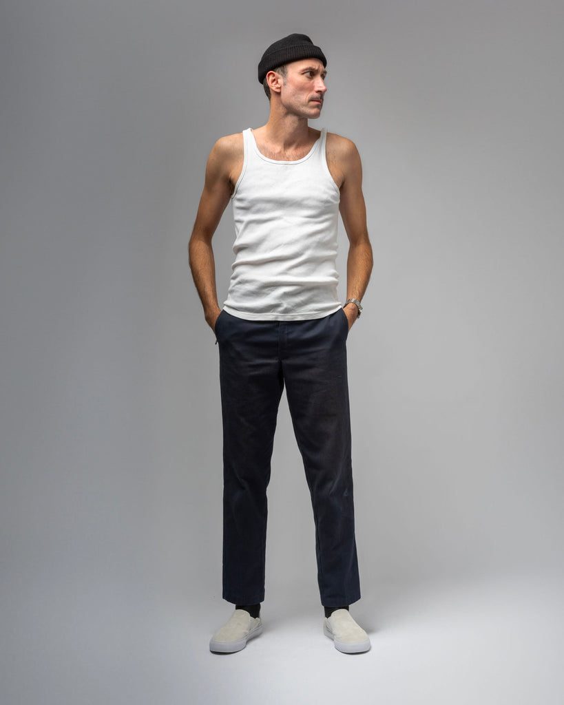 A man in a black beanie, white tank top, dark pants, and WARSAW LTD's STRANGER SLIP-ON | WHITE / WHITE stands with hands in pockets against a plain gray background, showcasing comfort and durability in his understated style.