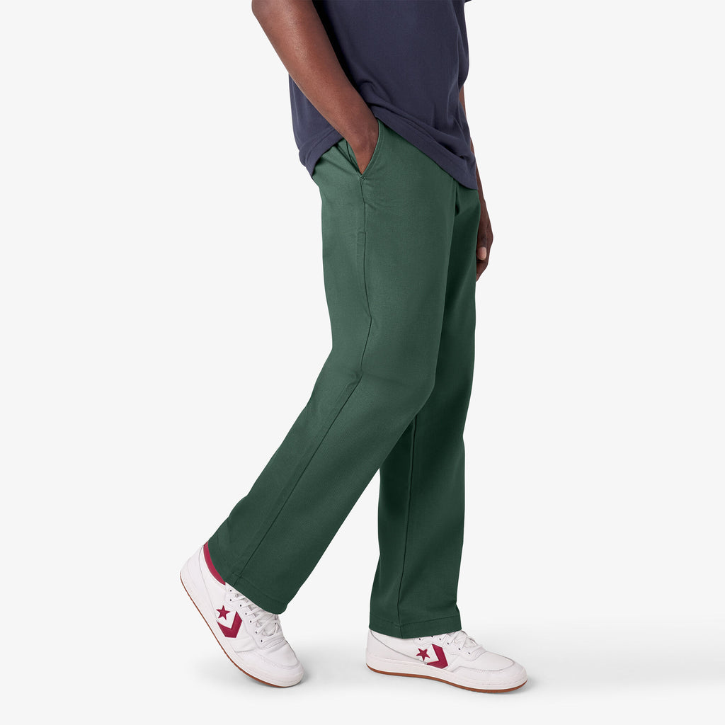 Side view of a person in DICKIES GUY MARIANO PANT PINE NEEDLE GREEN and white sneakers with pink details, focusing on the outfit from waist to feet.