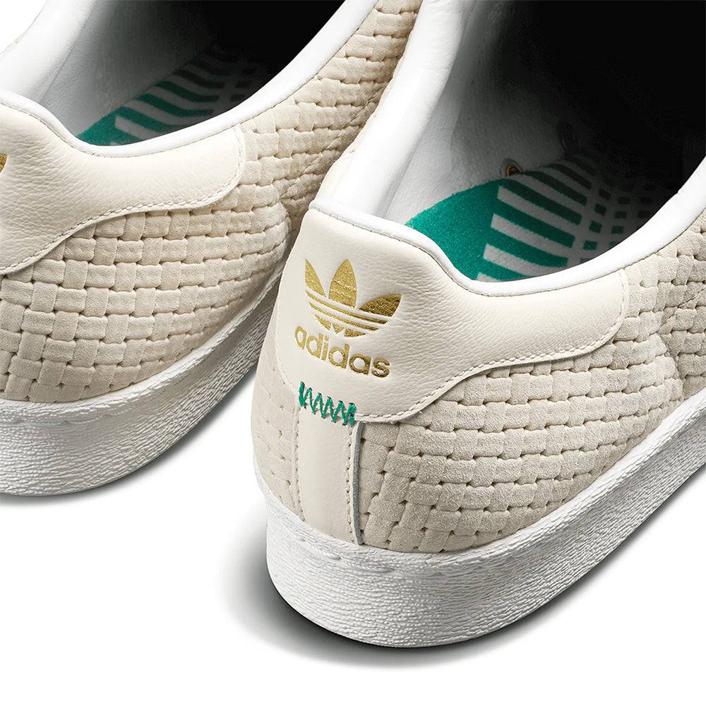 Close-up of the heel area on ADIDAS X ARROW & BEAST SUPERSTAR ADV WONDER WHITE / CRYSTAL WHITE, featuring a woven texture, gold adidas logo, green stitching, and a patterned green insole.