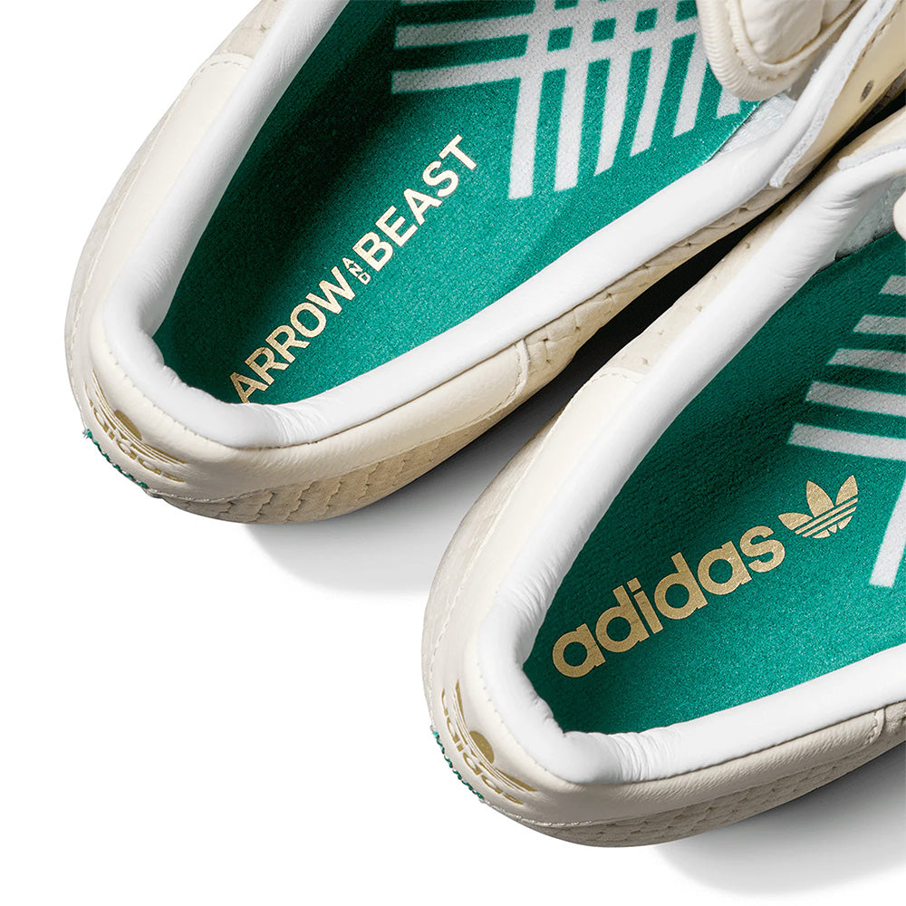 Close-up of two white ADIDAS X ARROW & BEAST SUPERSTAR ADV WONDER WHITE/CRYSTAL WHITE skate shoes, showing green insoles with "ARROW & BEAST" and "adidas" branding in white and gold, highlighting the premium cupsole construction.
