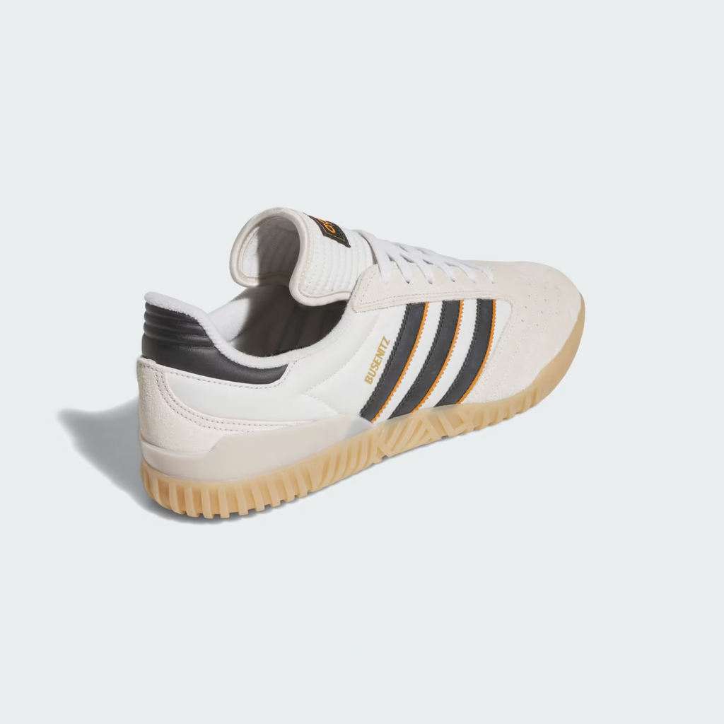 The ADIDAS BUSENITZ INDOOR SUPER in Bliss, Core Black, and Off White features a durable leather upper, gum sole, and gold "BUSENITZ" on the side. Shown from a rear side angle with black and orange stripes.