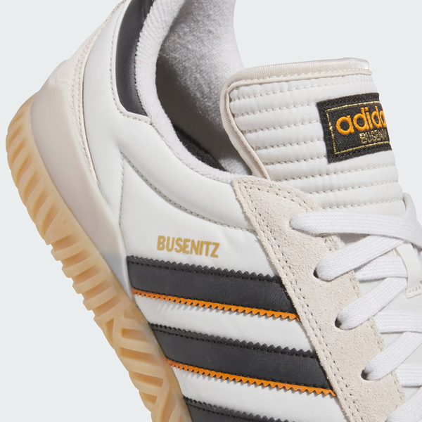 Close-up of the ADIDAS BUSENITZ INDOOR SUPER BLISS / CORE BLACK / OFF WHITE sneaker, showcasing black and orange stripes, gold branding, a gum sole, and a durable leather upper inspired by classic skateboarding shoes.
