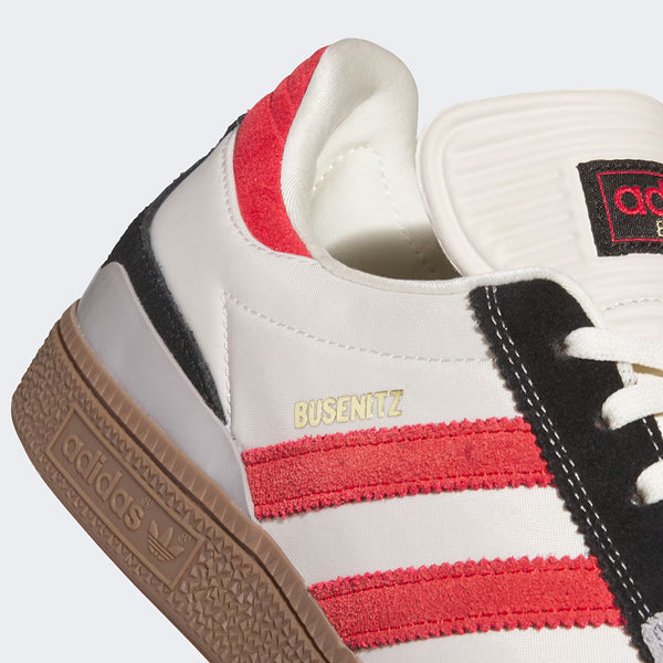 Close-up of the ADIDAS Busenitz Bliss / Better Scarlet / Gum shoe with red stripes, Adidas branding on the gum sole, gold "BUSENITZ" text, and a white and black upper.