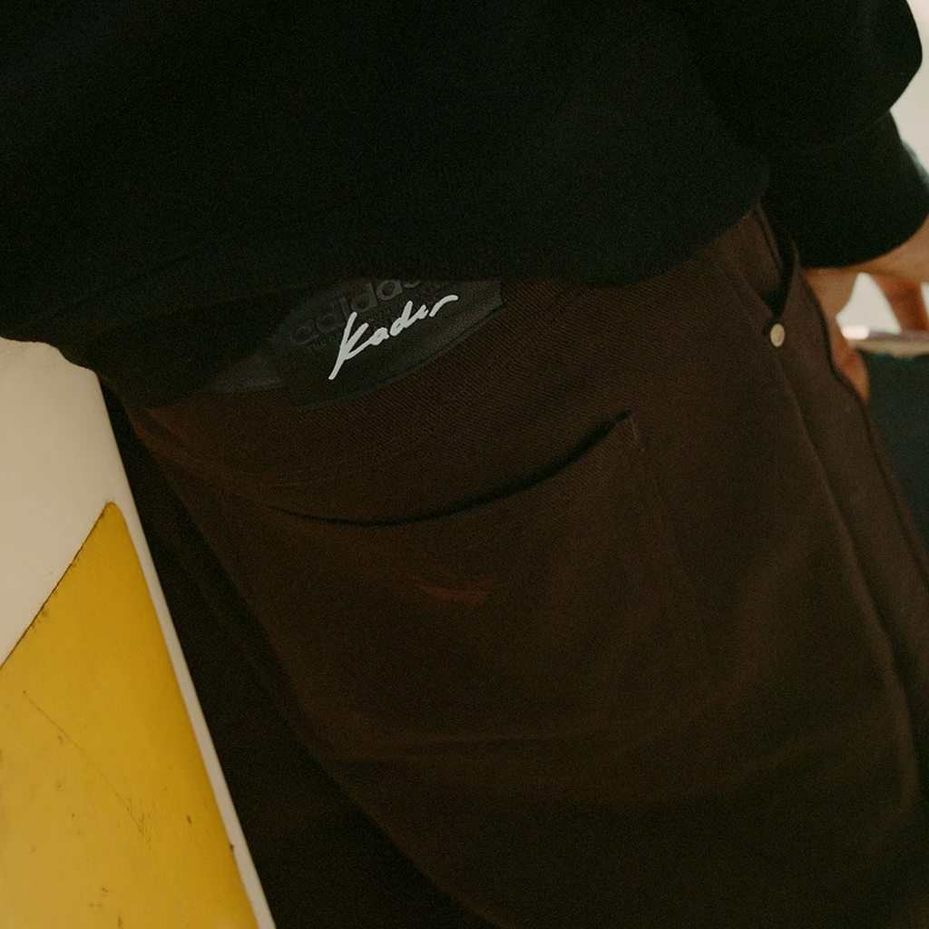 A close-up shot shows the back pocket of someone wearing dark brown, Baggy Fit ADIDAS SKATEBOARDING KADER PANTS from DGK, with a visible Kader-branded label above the pocket. The person is leaning against a yellow and white surface.