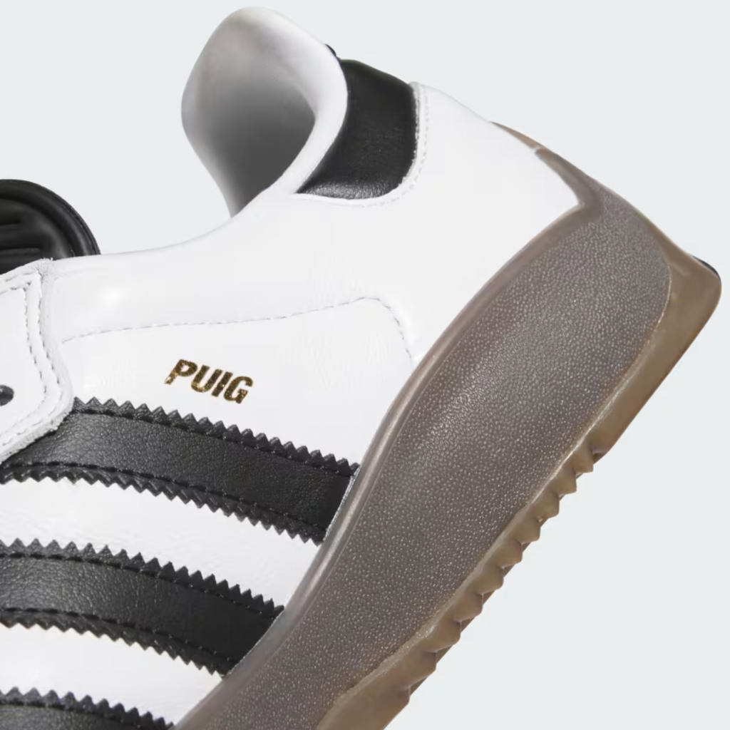 A close-up of the ADIDAS PUIG SAMBA ADV in Cloud White/Core Black/Gum, showcasing black stripes, the word "PUIG" in gold on the side, and Samba details such as a durable gum rubber outsole.