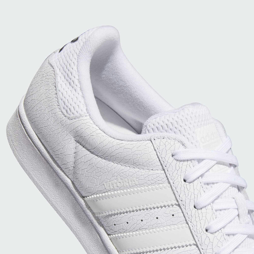 Close-up of the ADIDAS X VITORIA MENDONCA SUPERSTAR ADV CORE WHITE / WHITE REFLECTIVE sneaker, showcasing its textured leather and suede upper with three stripes on the side, a patterned sole, and a flexible cupsole.